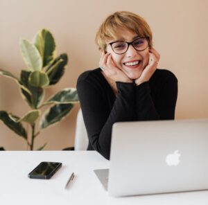 Cheerful woman in eyeglasses and black sweater smiling while working at a desk with a laptop indoors.